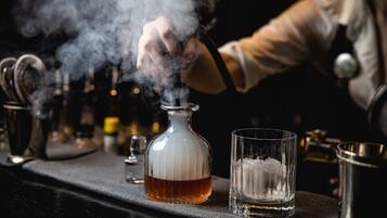 A bar setting with a glass decanter, a glass with a candle, and a metal shaker on a bar counter.