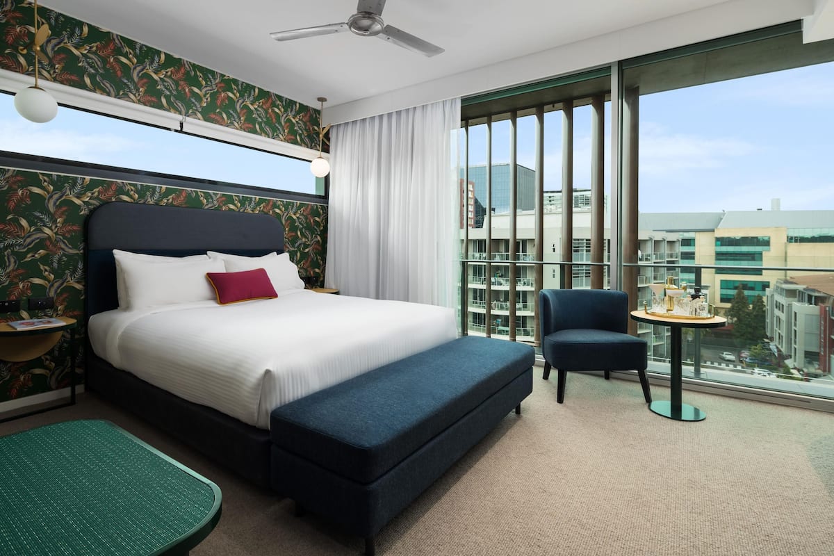 Habitación de hotel con una cama grande, una mesita, una silla y vistas a la ciudad a través de ventanas amplias.