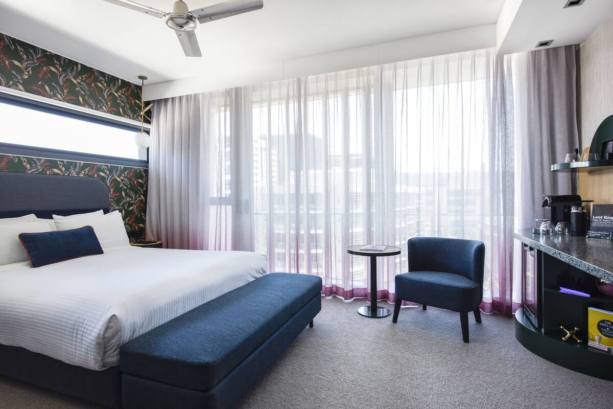 Habitación de hotel con cama, un banco, una silla, una mesita y vistas a la ciudad a través de cortinas translúcidas.