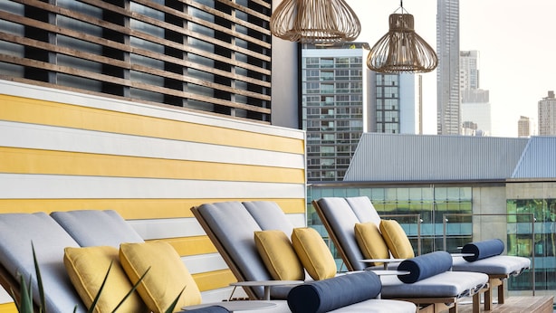 A rooftop lounge area with sun loungers, wooden flooring, and hanging wicker lamps.