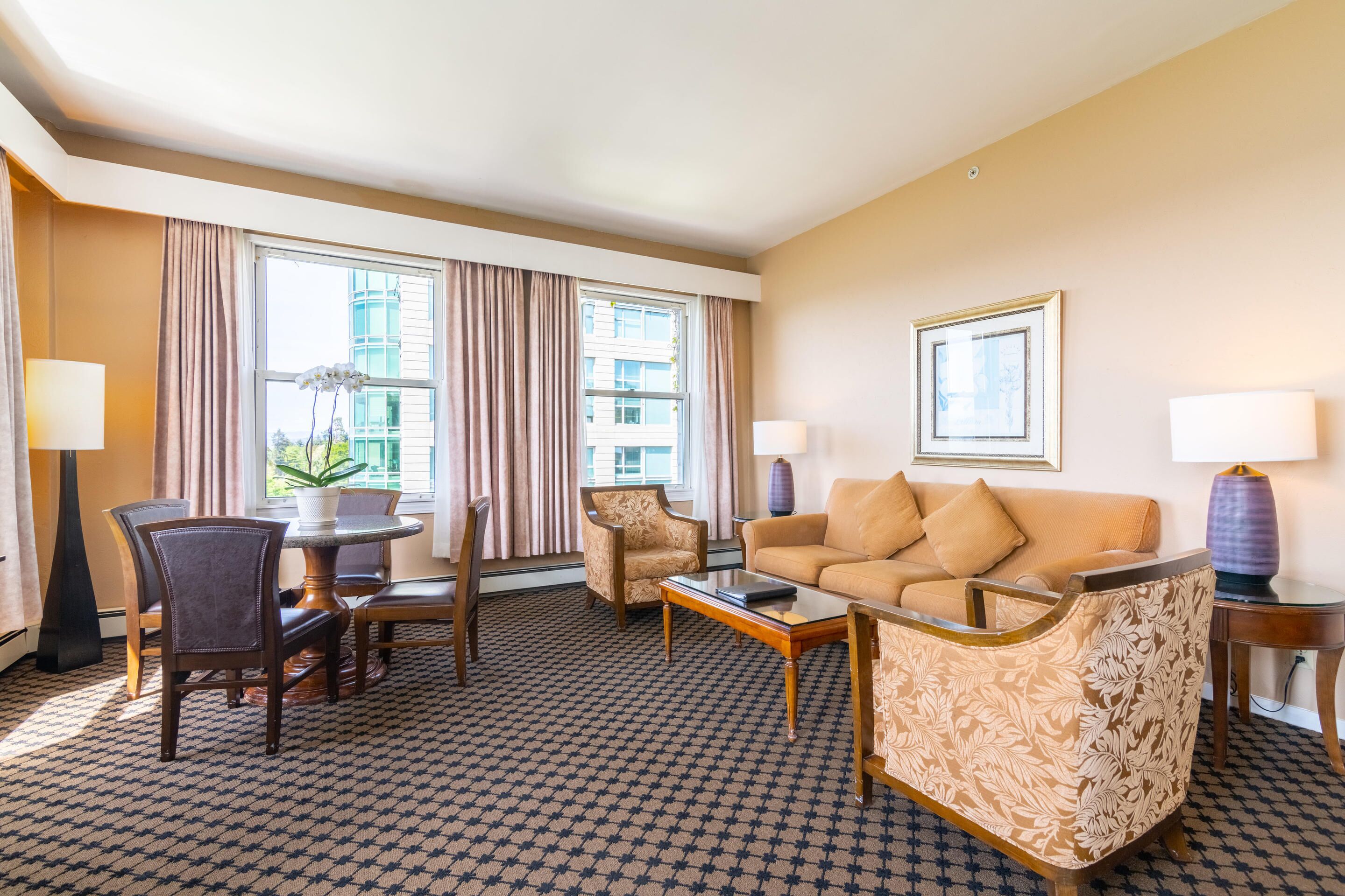 Habitación de hotel con sofá, sillón, mesa de comedor y sillas. Dos ventanas con cortinas, una lámpara y un cuadro en la pared.