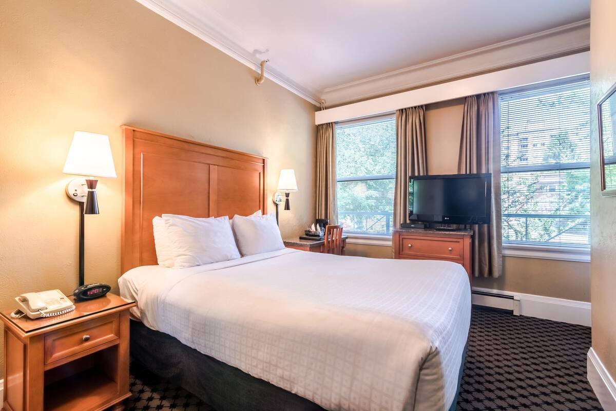 A hotel room with a large bed, two bedside lamps, a TV, a nightstand with a phone, and a window with curtains.