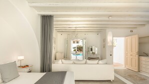 A modern hotel room with a bed, a sofa, a desk with a lamp, a mirror, and a view of the pool. - Corfos Hotel (Mykonos)