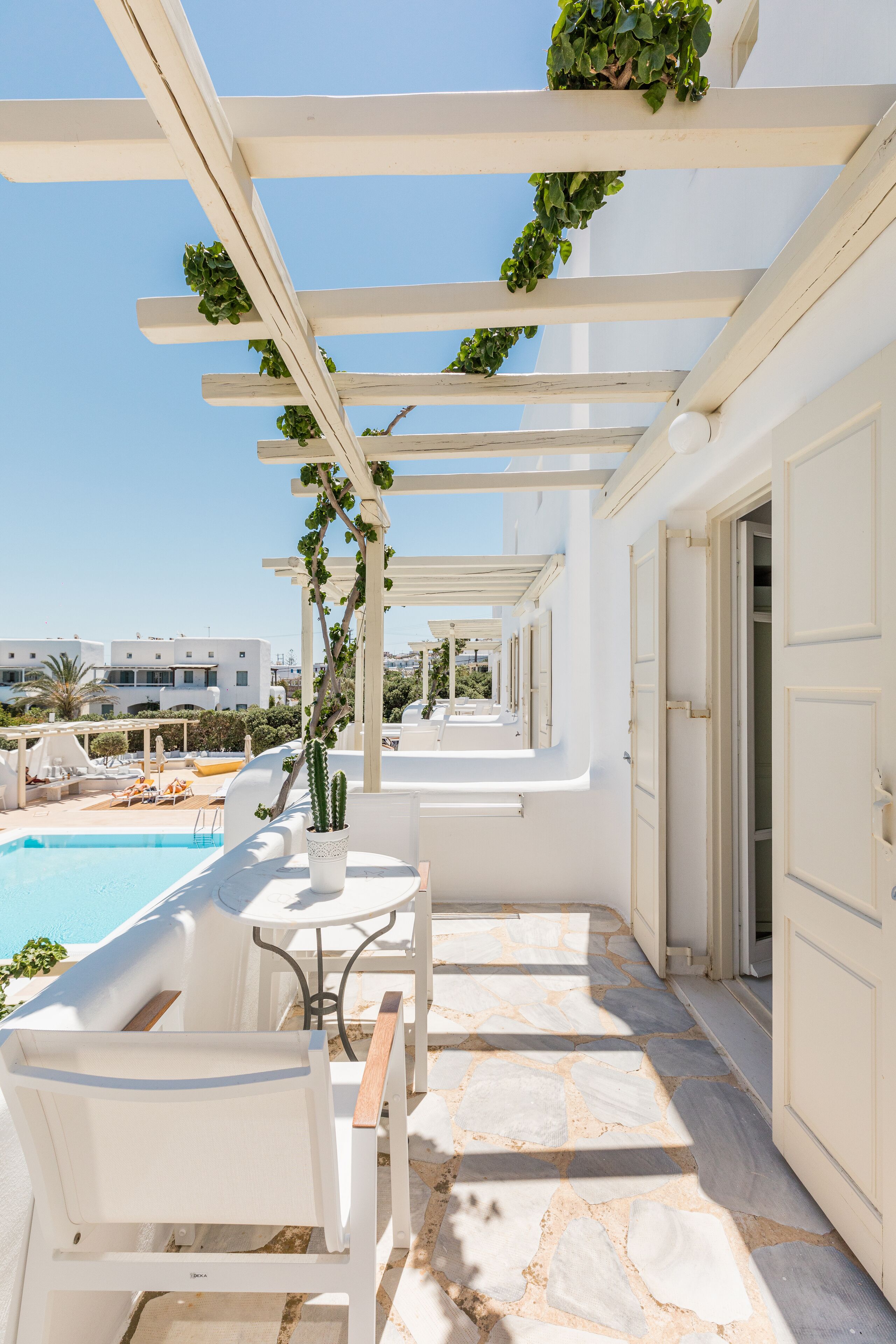 A white outdoor seating area with a pool, stone flooring, and a pergola with greenery.