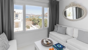 A modern living room with a white sofa, a coffee table with a bowl of oranges, and a view of the sea through large windows. - Corfos Hotel (Mykonos)