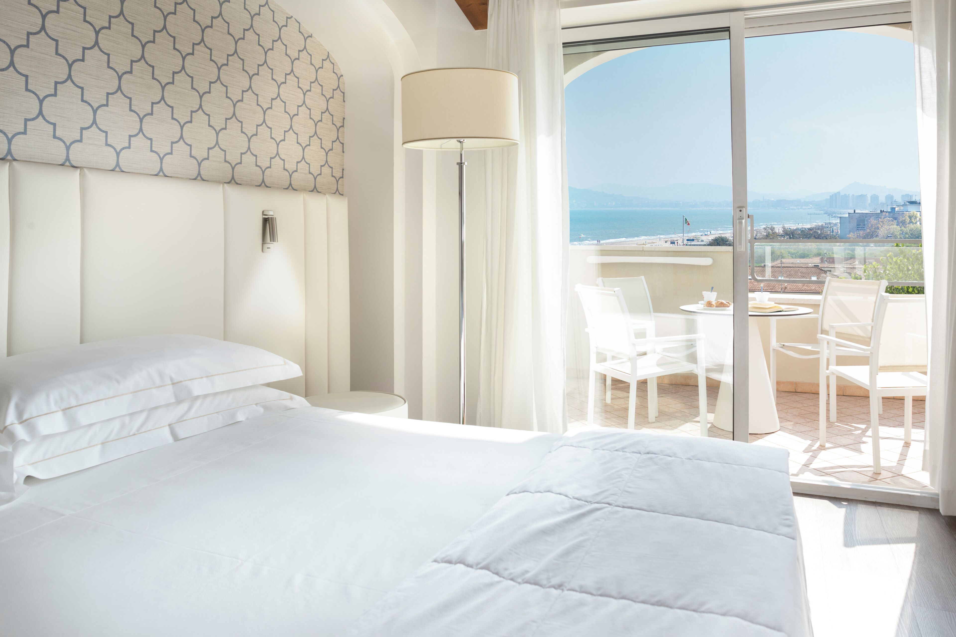 A hotel room with a bed, a floor lamp, a balcony with a table and chairs, and a view of the sea.