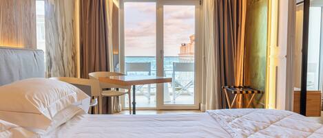 A hotel room with a bed, a chair, a table, and a view of the sea through a glass door.