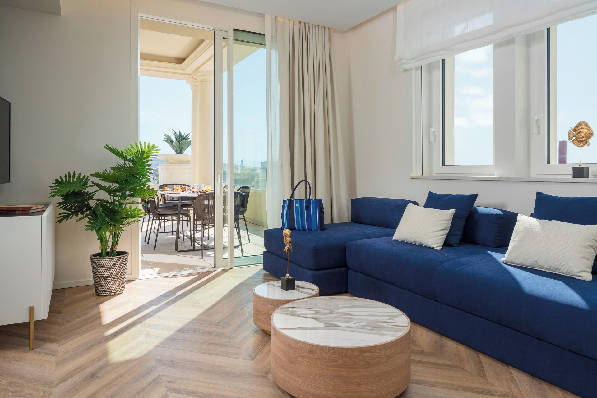 A modern living room with a blue sofa, a round coffee table, a dining area with a table and chairs, and a balcony with a view.