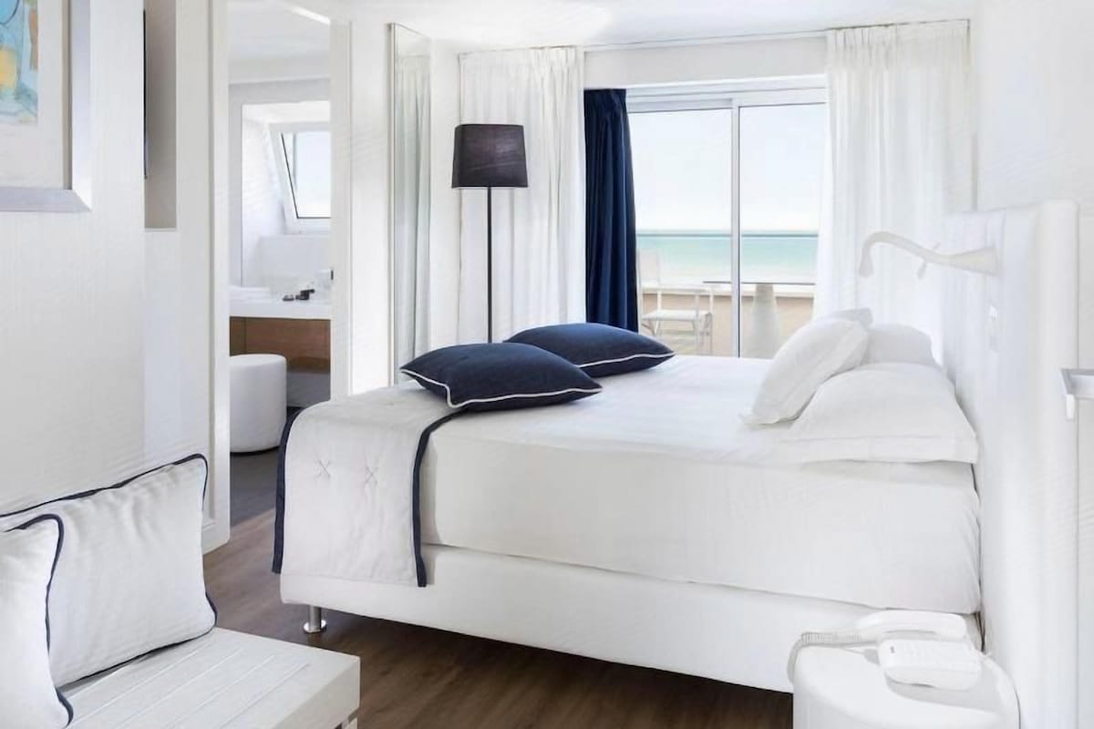 A hotel room with a bed, a bedside table, a lamp, and a view of the beach.