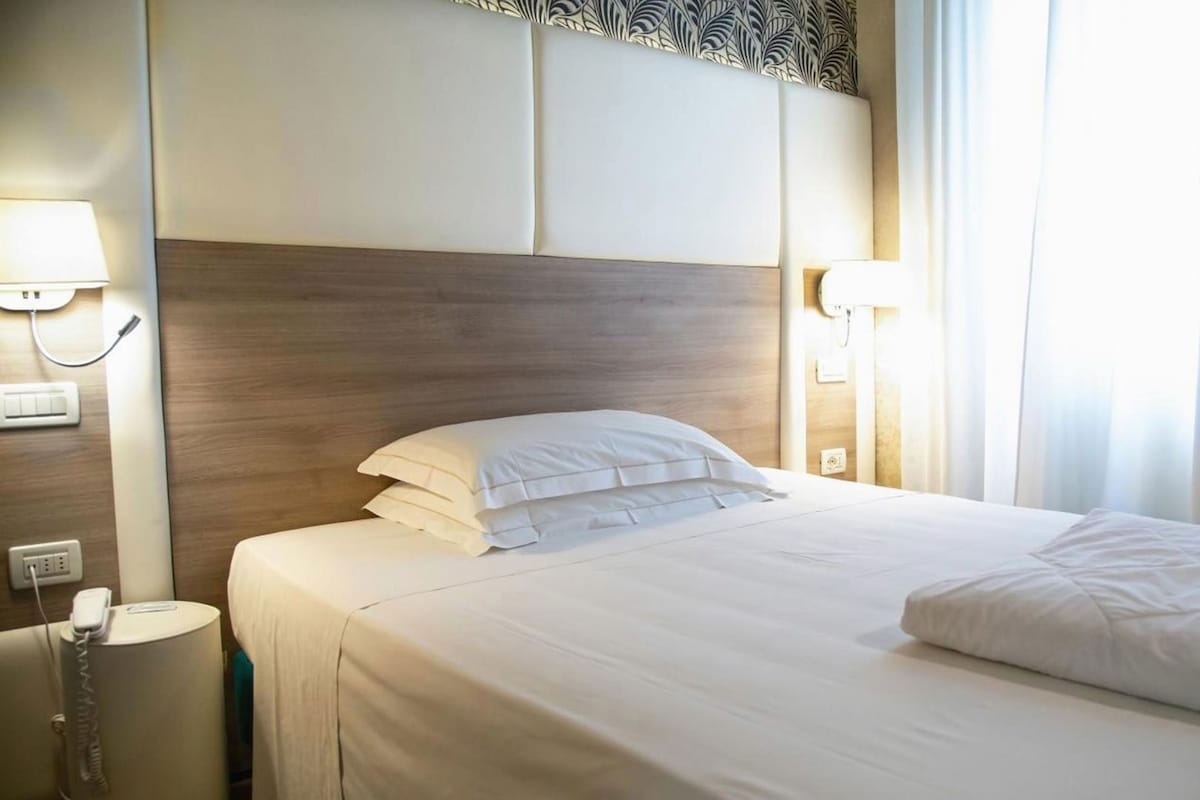 A hotel room with a neatly made bed, bedside lamps, a telephone, and a power outlet.
