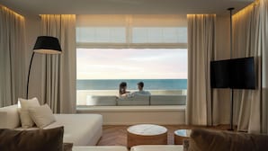 A modern living room with a large window overlooking the sea, a sofa, a coffee table, and a TV. - Hotel Corallo (Riccione)