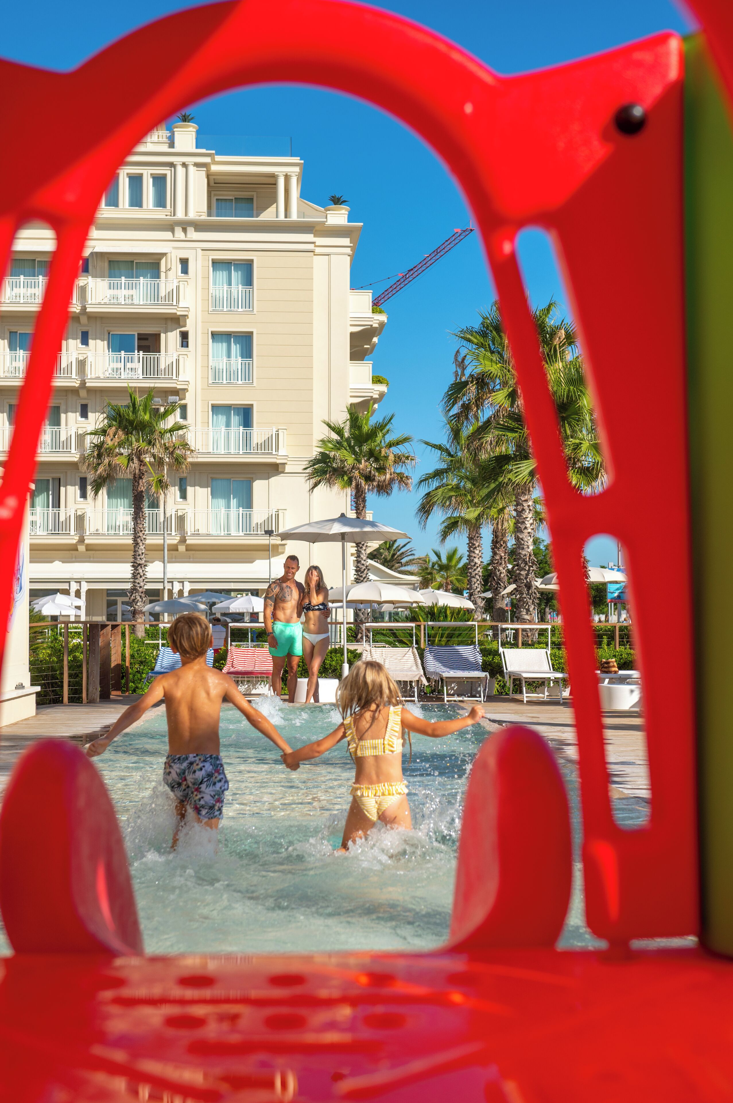 A swimming pool area with a building in the background, palm trees, and lounge chairs.