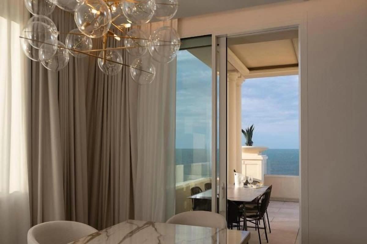 A dining area with a marble table and chairs, a chandelier, and a view of the sea through large windows.