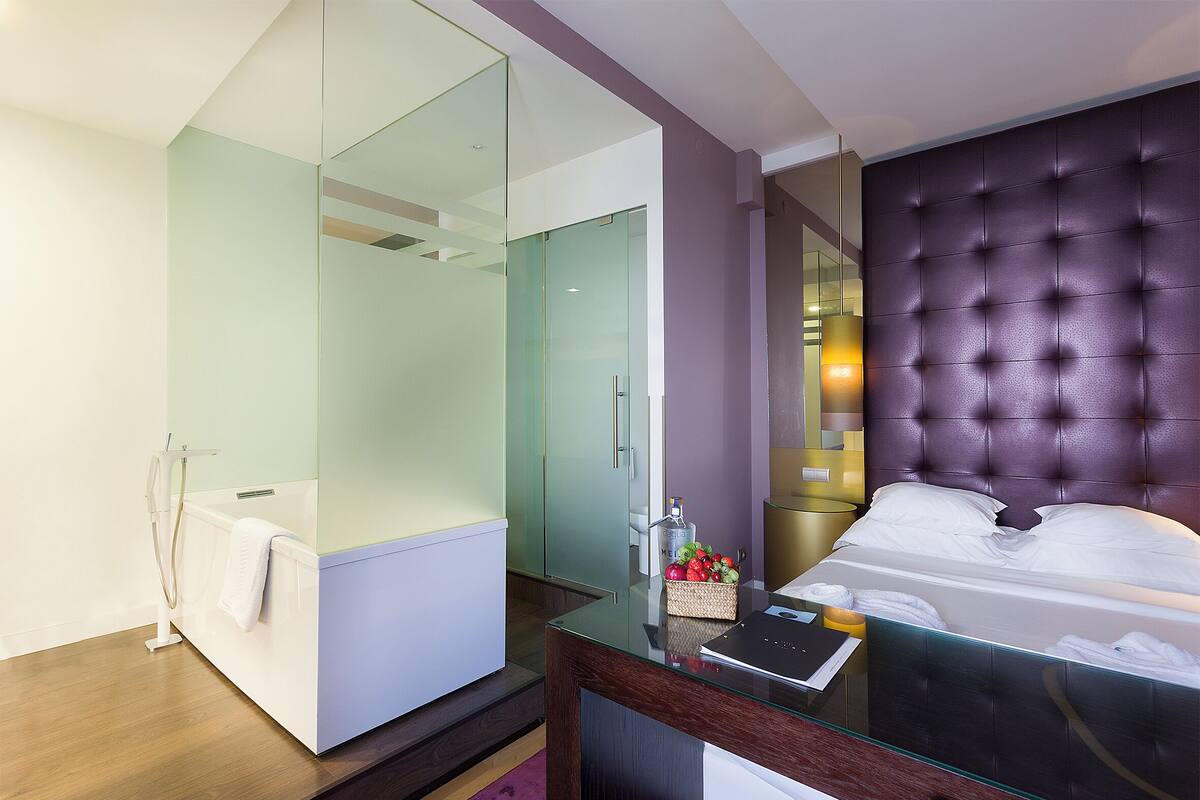 A modern hotel room with a glass shower enclosure, a tufted headboard, and a bedside table with a lamp.