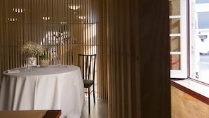 Un comedor con una mesa redonda dispuesta para dos personas, una silla, una planta en maceta y una ventana con vistas al exterior.