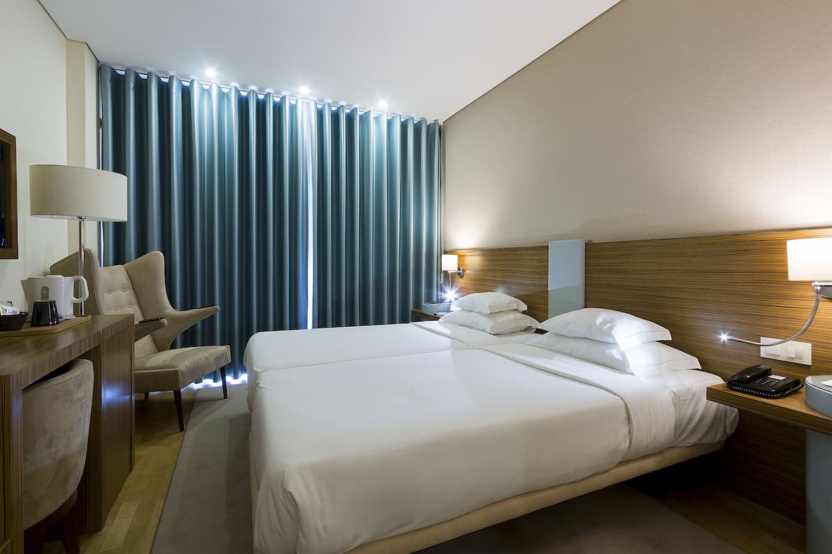 A hotel room with a large bed, a desk with a lamp, a chair, and a nightstand with a phone and lamp.
