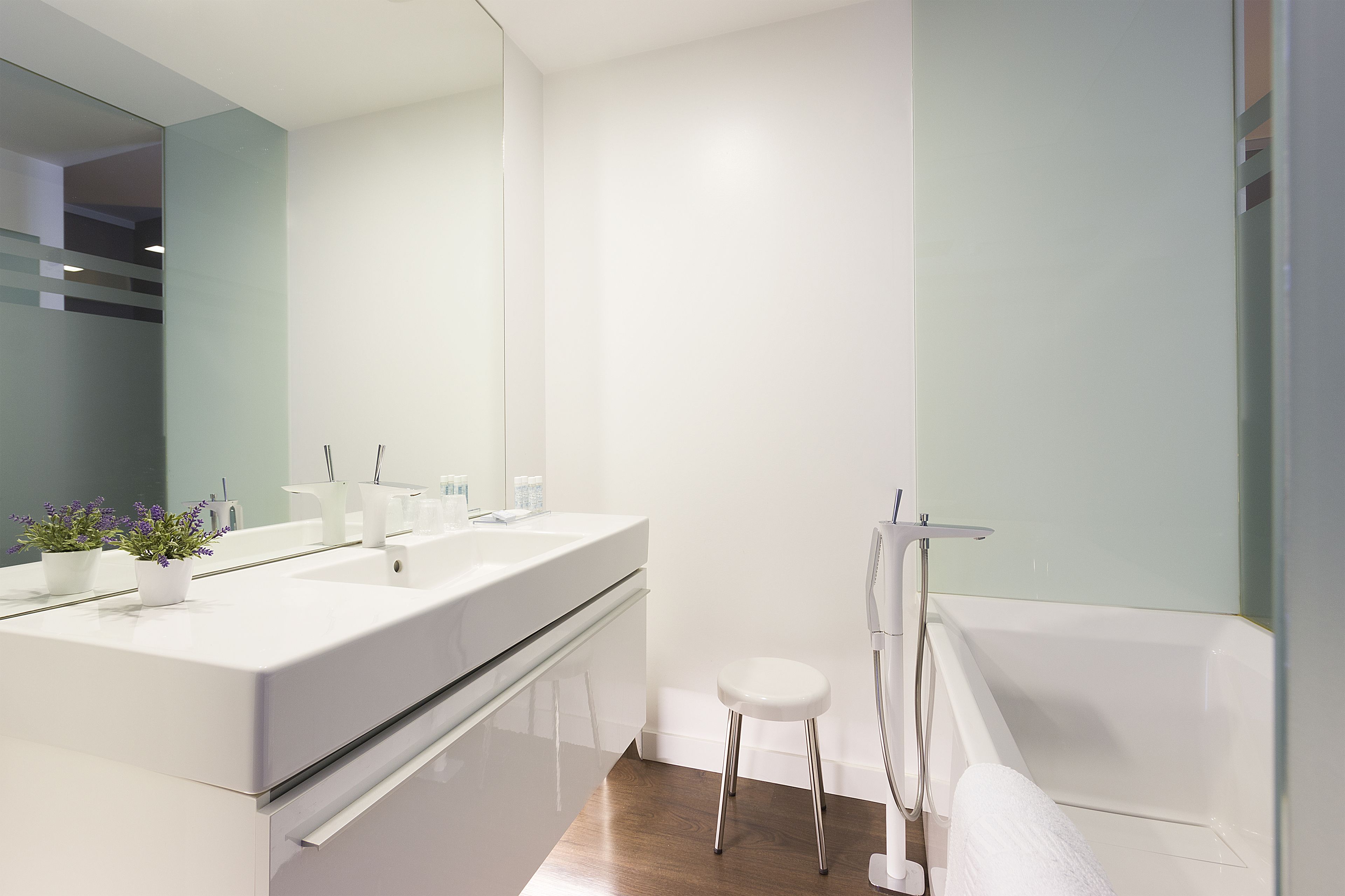 Un baño moderno con un espejo grande, dos lavamanos, una bañera blanca y un taburete pequeño.