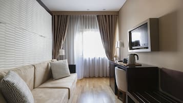 A hotel room with a sofa, a TV, a desk with a lamp, and a window with curtains.