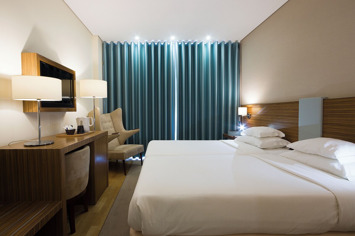 A hotel room with a large bed, a desk with a chair, a TV, and a nightstand with a lamp.