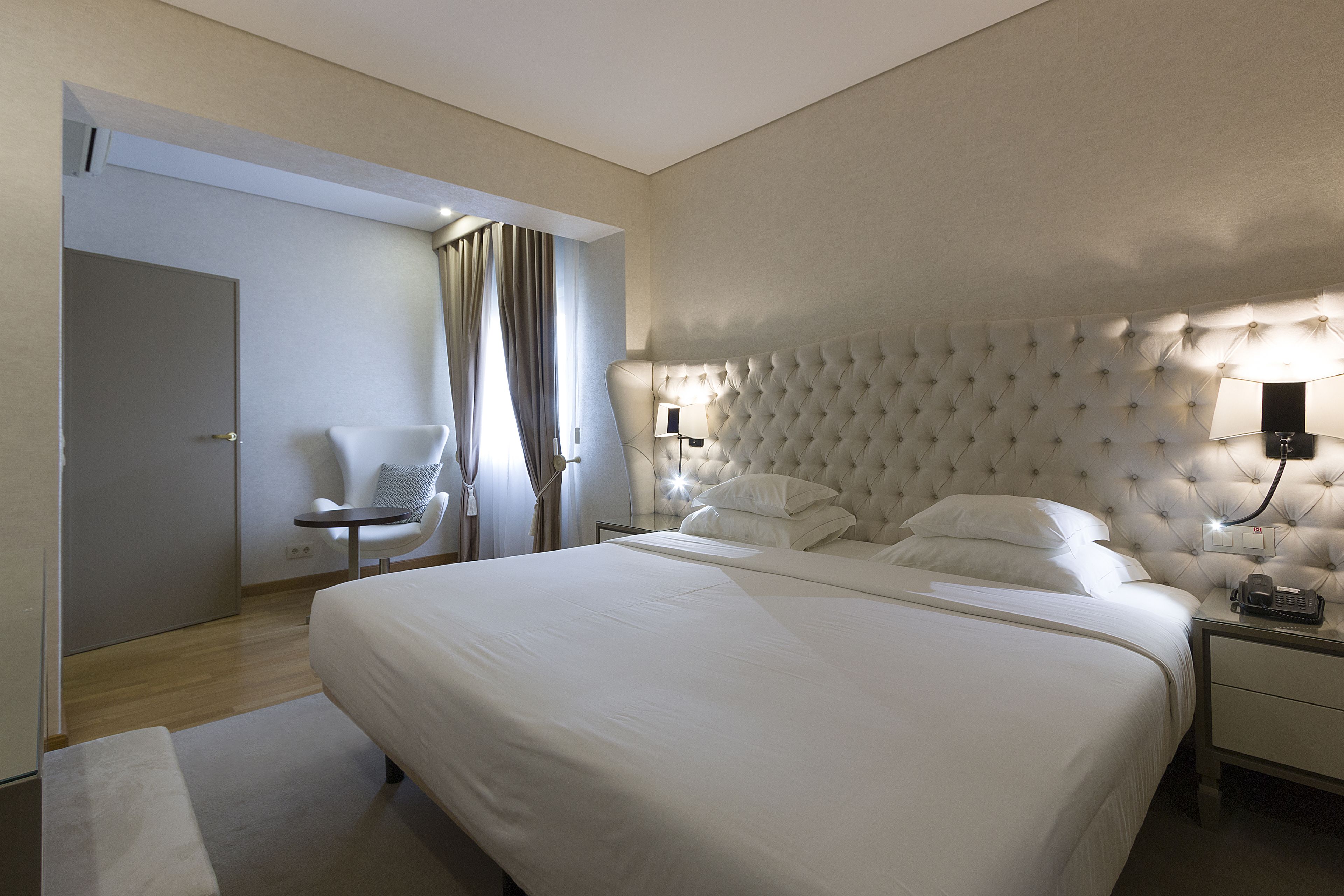 Una habitación de hotel moderna con una cama grande, una mesita de noche con lámpara, una silla y una puerta.