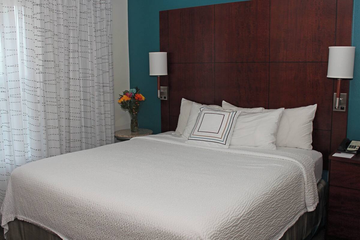 A neatly made bed with a white comforter and pillows, a wooden headboard, two bedside lamps, and a nightstand with a vase of flowers.