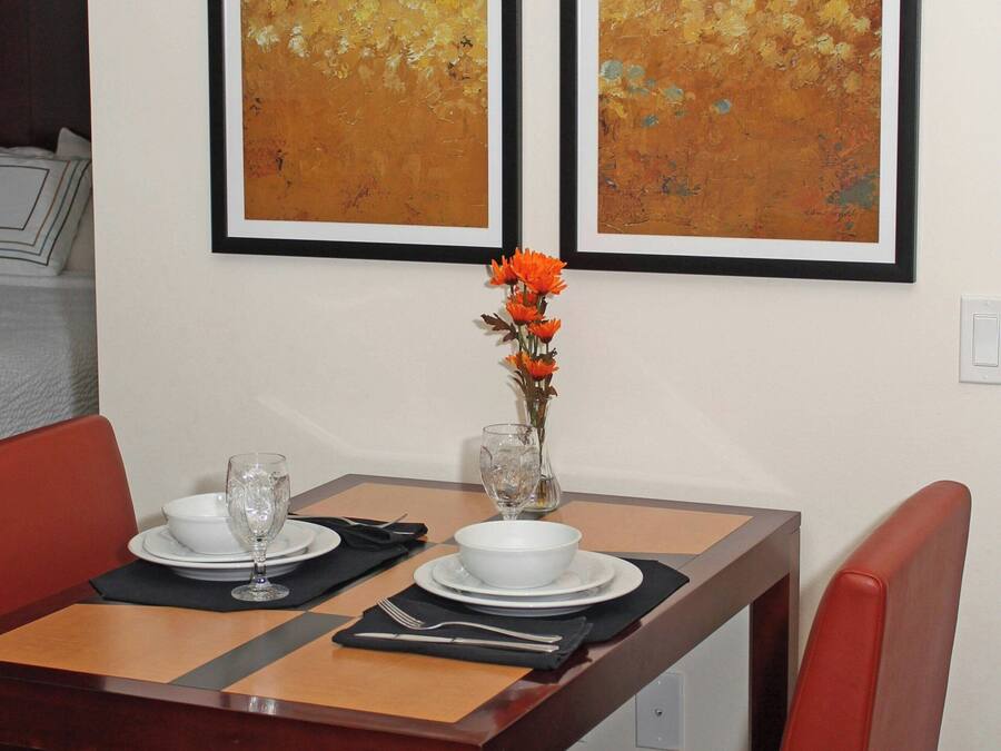 A dining table set with white plates, glasses, and a vase with orange flowers. Two abstract paintings hang on the wall above the table.