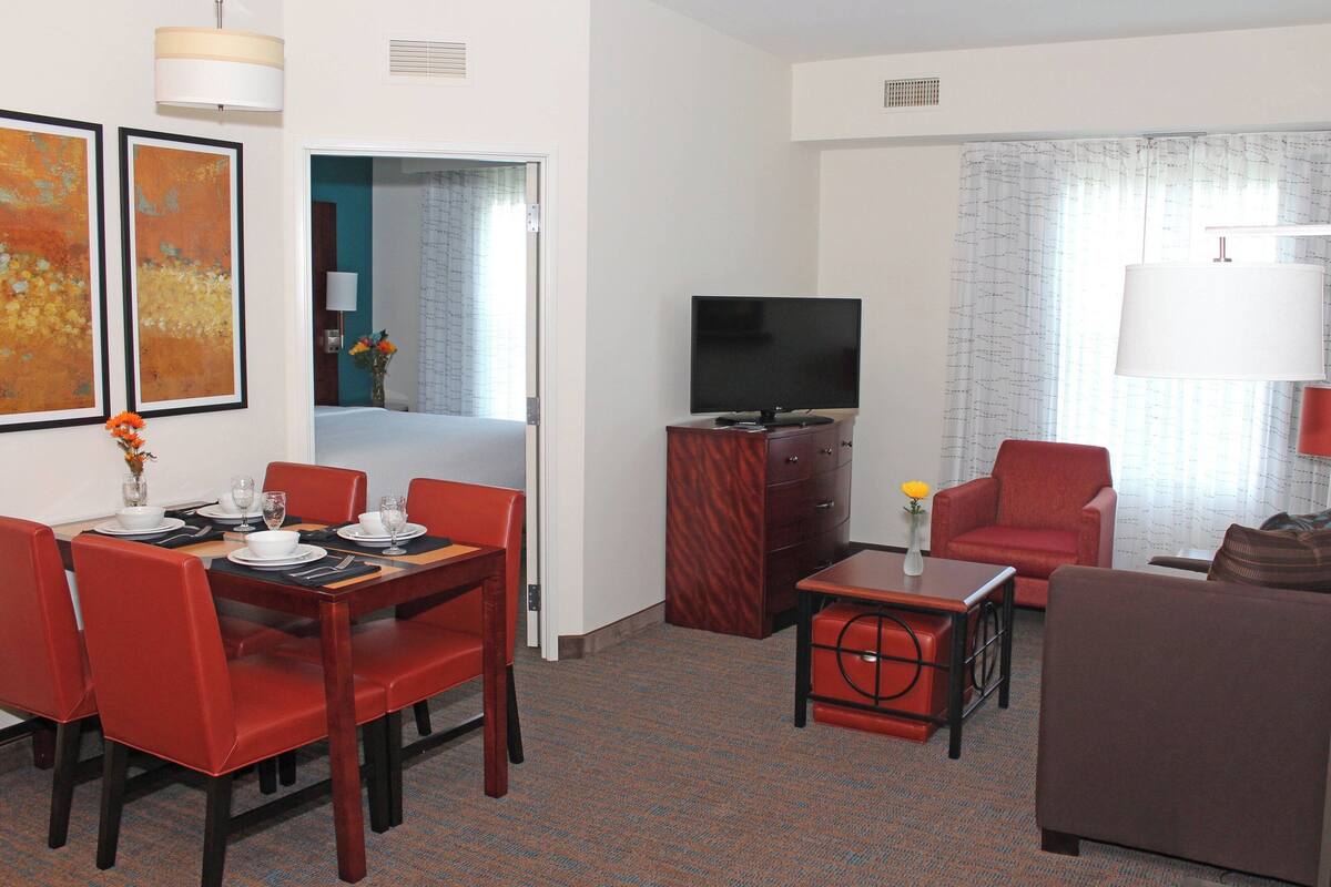 A hotel room with a dining area, a TV, a sofa, and a small table with a vase of flowers.
