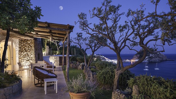 A patio with outdoor seating, a stone wall, and a view of the sea at night.