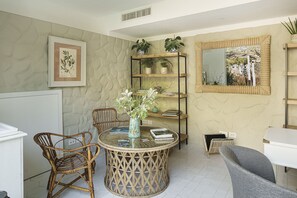 Una habitación con una mesa redonda de mimbre, dos sillas de mimbre, un armario blanco, una estantería con plantas y un espejo grande en la pared.