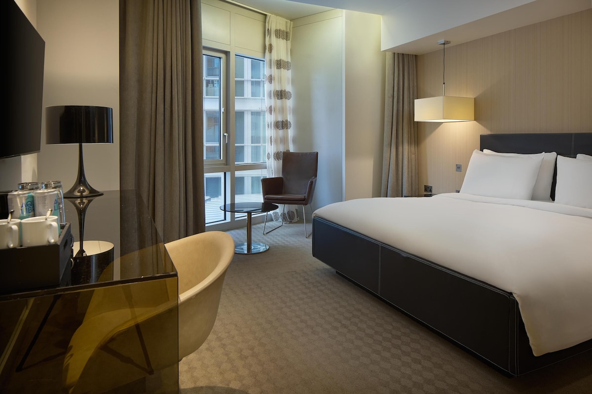 A hotel room with a large bed, a desk with a lamp, a chair, and a view of the outdoors.
