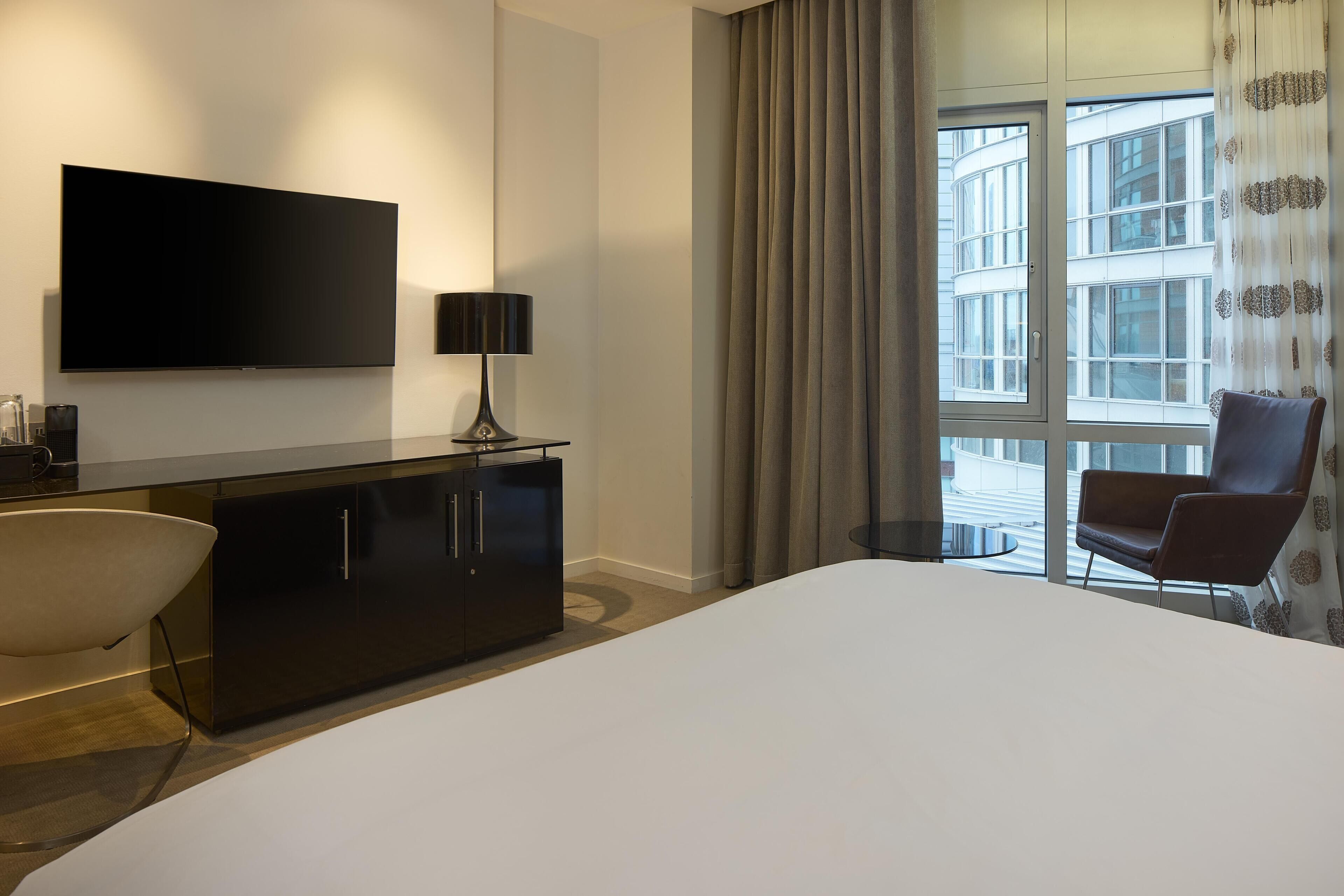 Ein Hotelzimmer mit einem großen Bett, einem flachbildfernseher an der Wand, einem modernen Schreibtisch mit Lampe, einem Sessel und Blick auf die Stadt durch große Fenster.