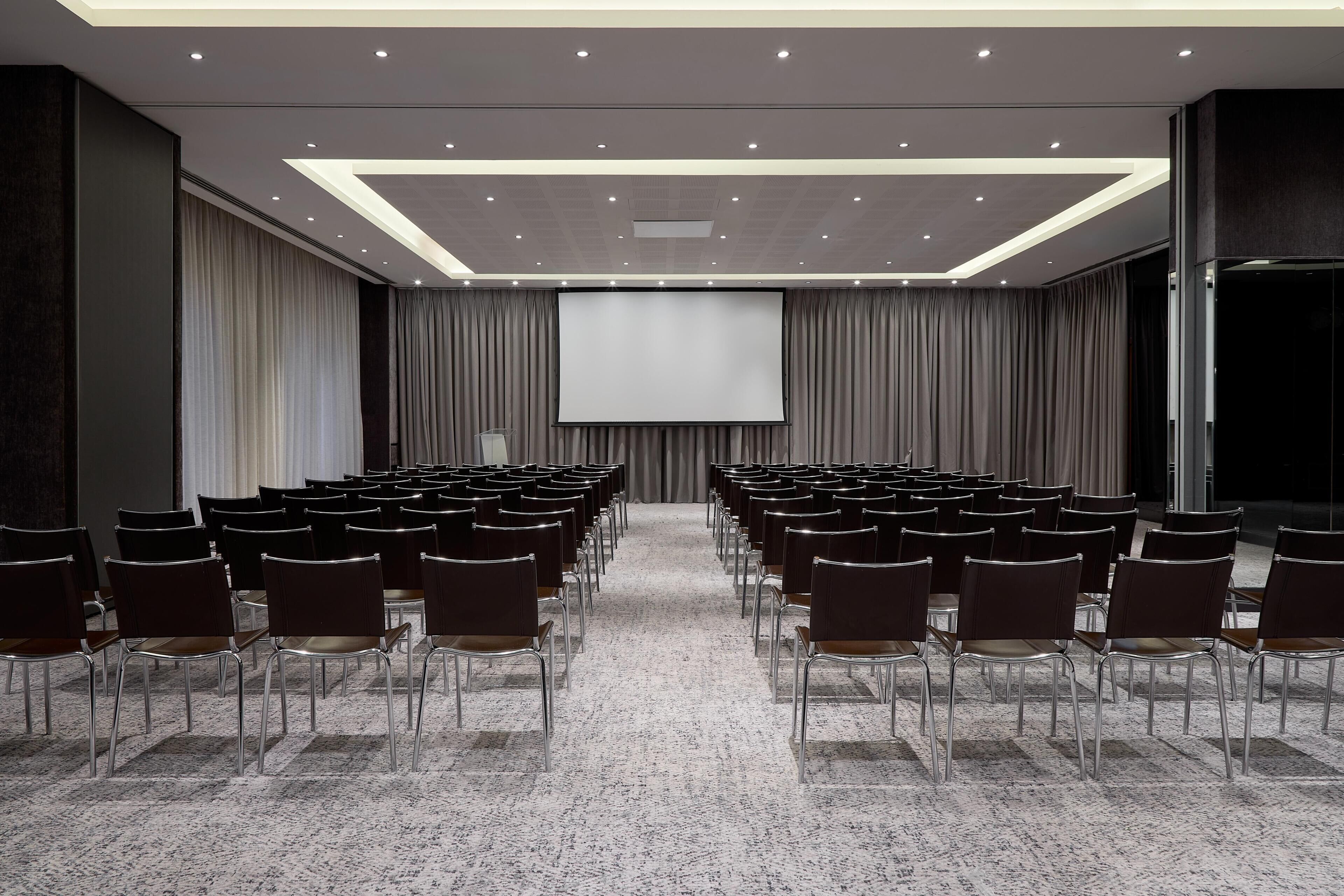 A conference room with rows of chairs facing a large screen, a carpeted floor, and a modern ceiling design.