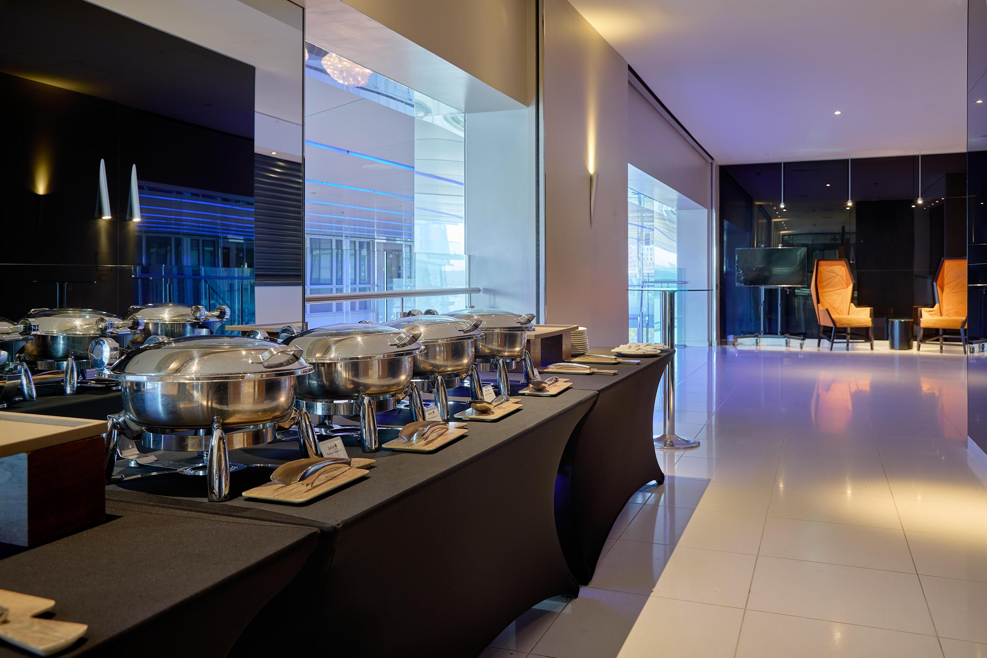 Ein modernes Buffet mit mehreren Edelstahl-Servierstationen, einer langen Theke und einer Sitzecke mit orangefarbenen Stühlen.
