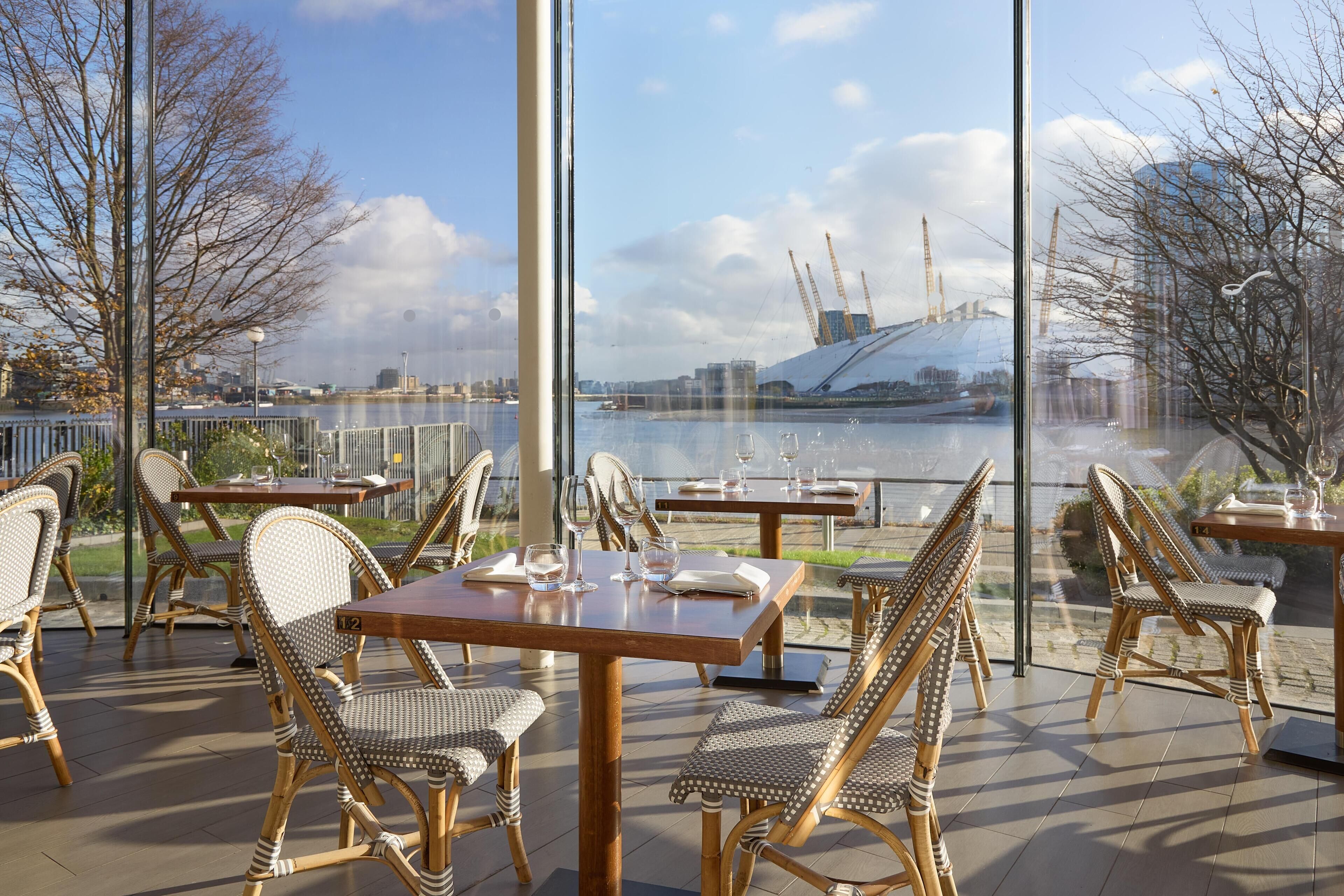 An outdoor dining area with wicker chairs and tables, overlooking a river with a bridge and buildings in the background.