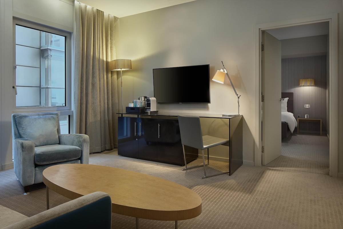 A modern hotel room with a flat-screen TV mounted on the wall, a desk with a chair, a sofa, and a large window with curtains.