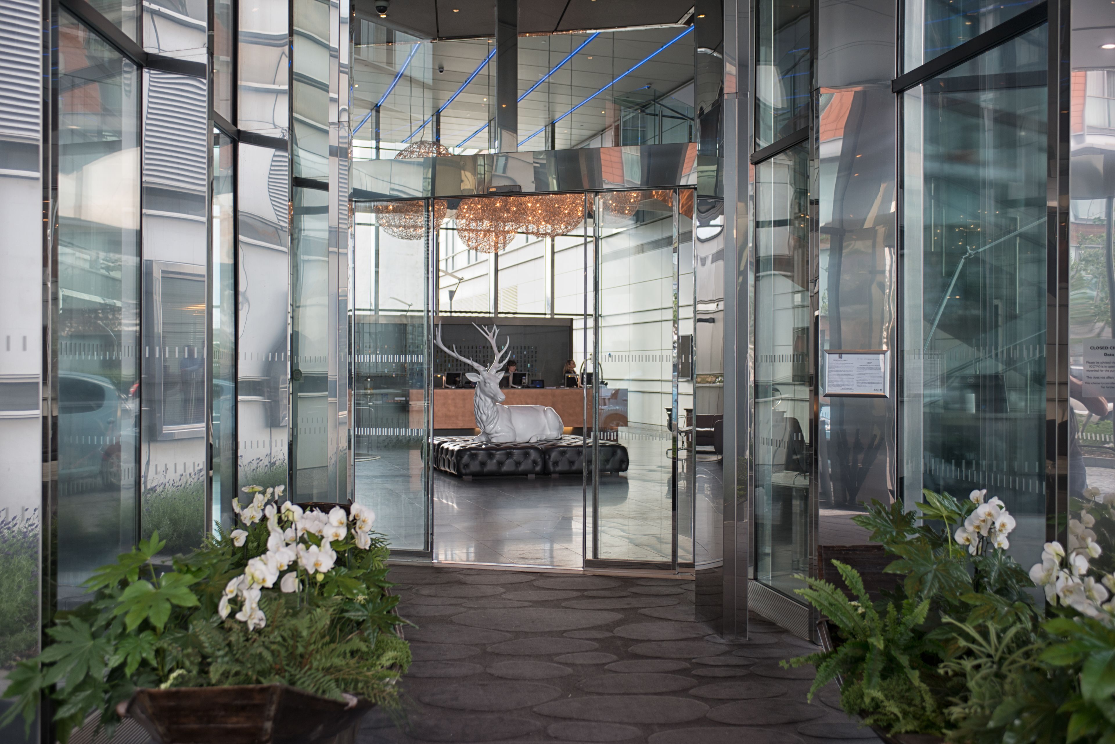 Un hall d’hôtel moderne avec des murs en verre, un espace salon central et un lustre.