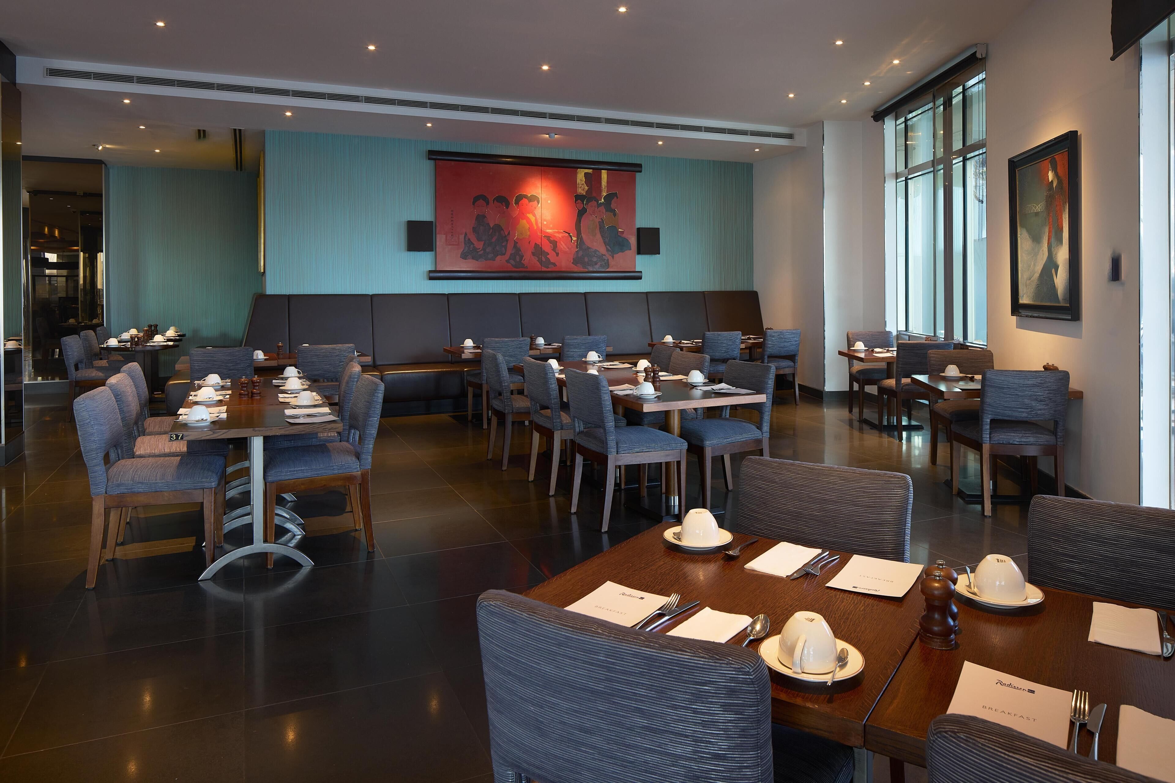 Un restaurant doté de nombreuses tables et chaises, d’un grand tableau accroché au mur et offrant une vue sur l’extérieur grâce à de grandes fenêtres.