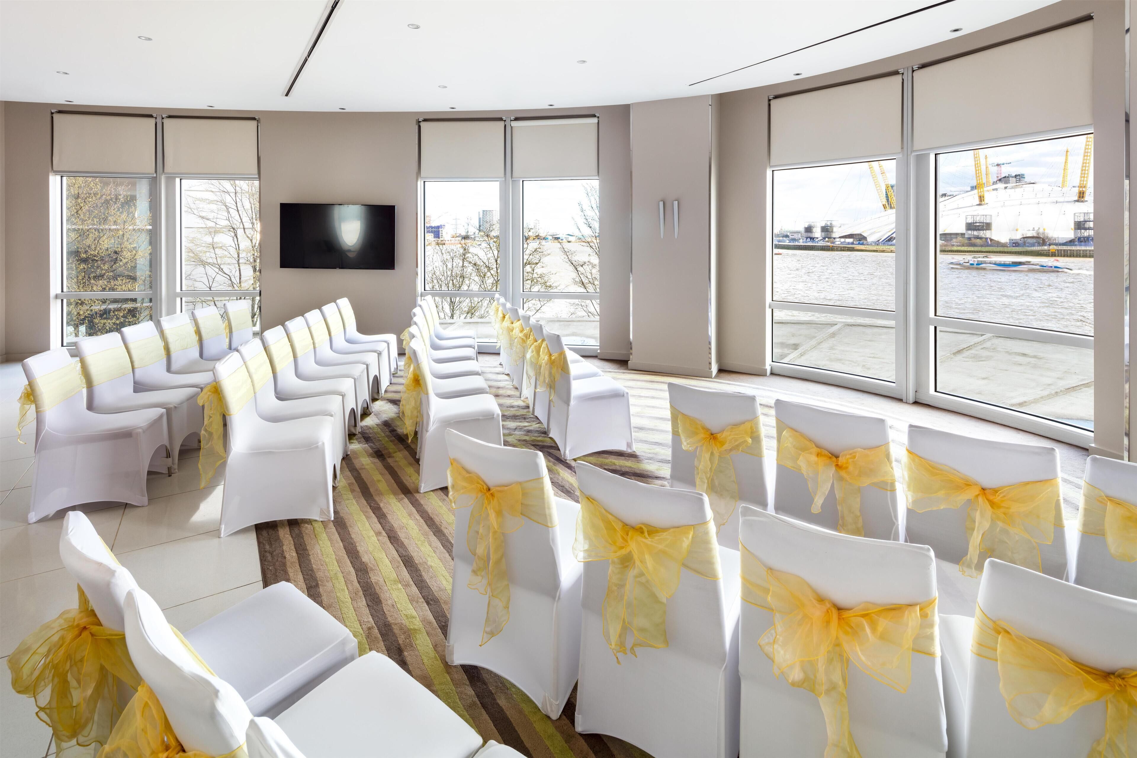 A conference room with rows of white chairs, yellow sashes, and a large window with a view of a construction site.