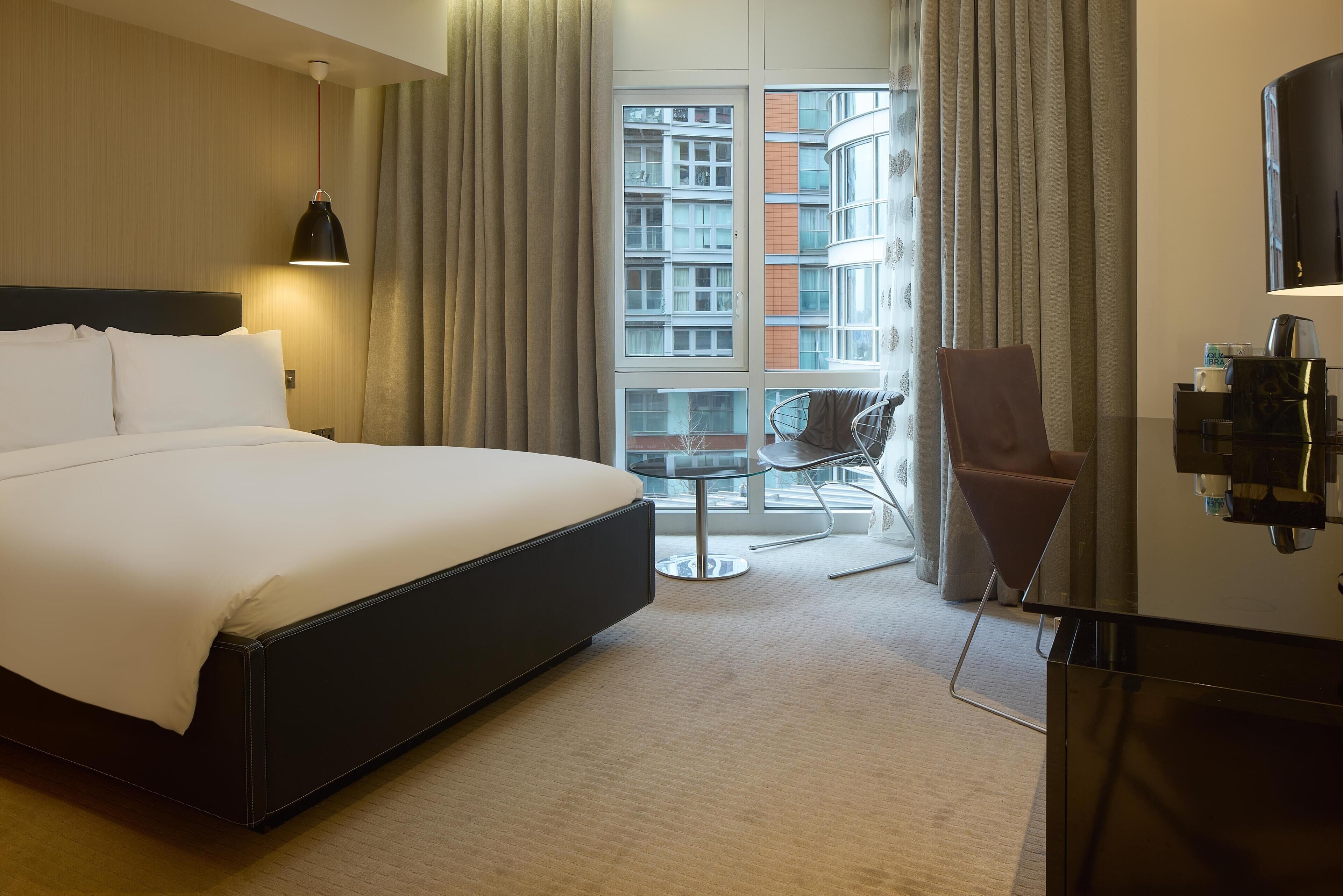 A hotel room with a large bed, a desk, a chair, a TV, and a view of a building through the window.