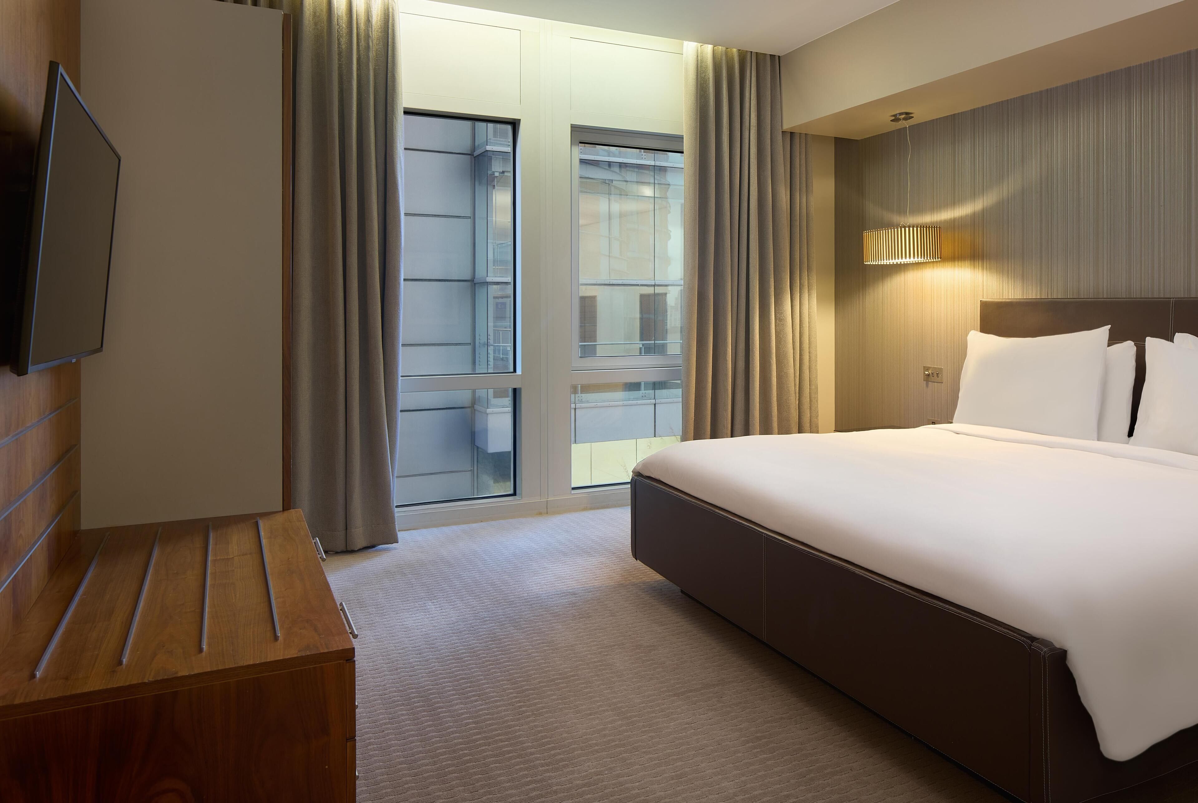 A hotel room with a large bed, a wooden dresser, a flat-screen TV, and a sliding glass door leading to a balcony.