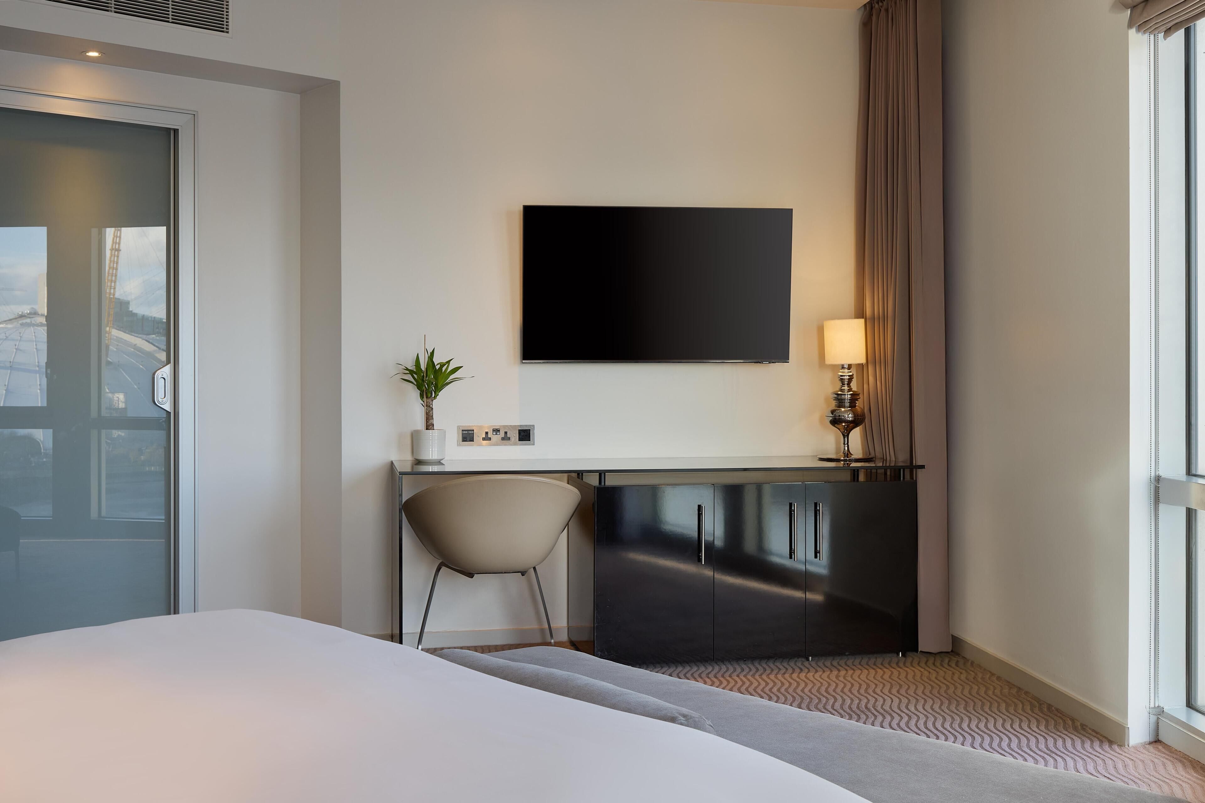 A hotel room with a bed, a desk with a chair, a flat-screen TV, a lamp, and a window with curtains.