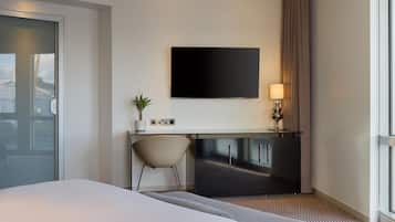 A hotel room with a bed, a desk with a chair, a flat-screen TV, a lamp, and a window with curtains.