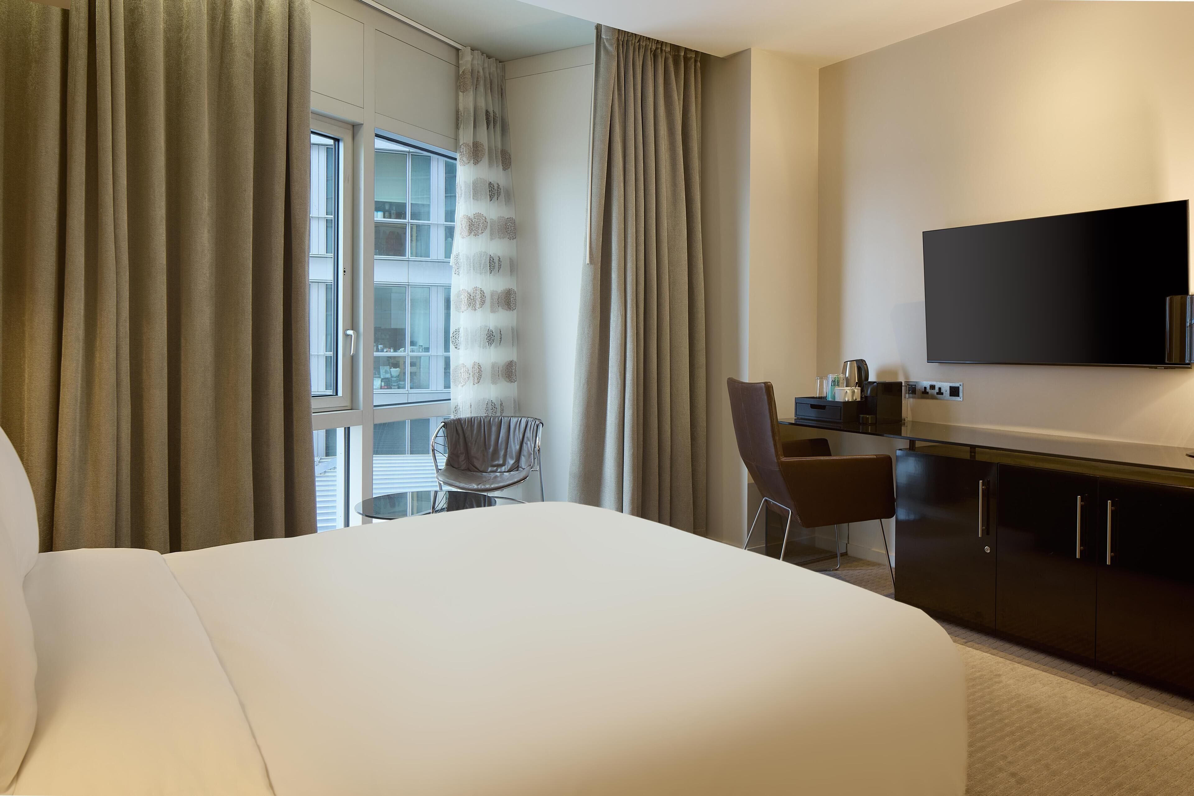 Ein Hotelzimmer mit einem großen Bett, einem Schreibtisch mit Stuhl, einem Flachbildfernseher und einem Fenster mit Vorhängen.