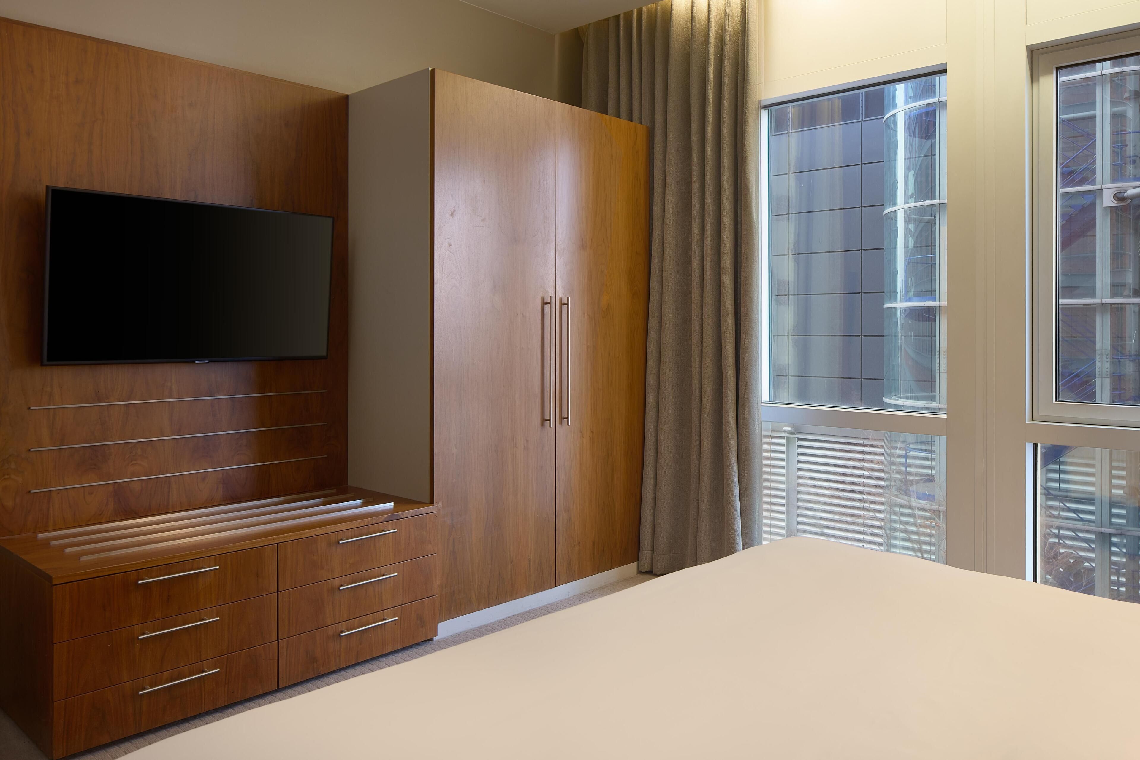 Une chambre avec une armoire en bois, un téléviseur à écran plat fixé au mur et une grande fenêtre avec des stores.