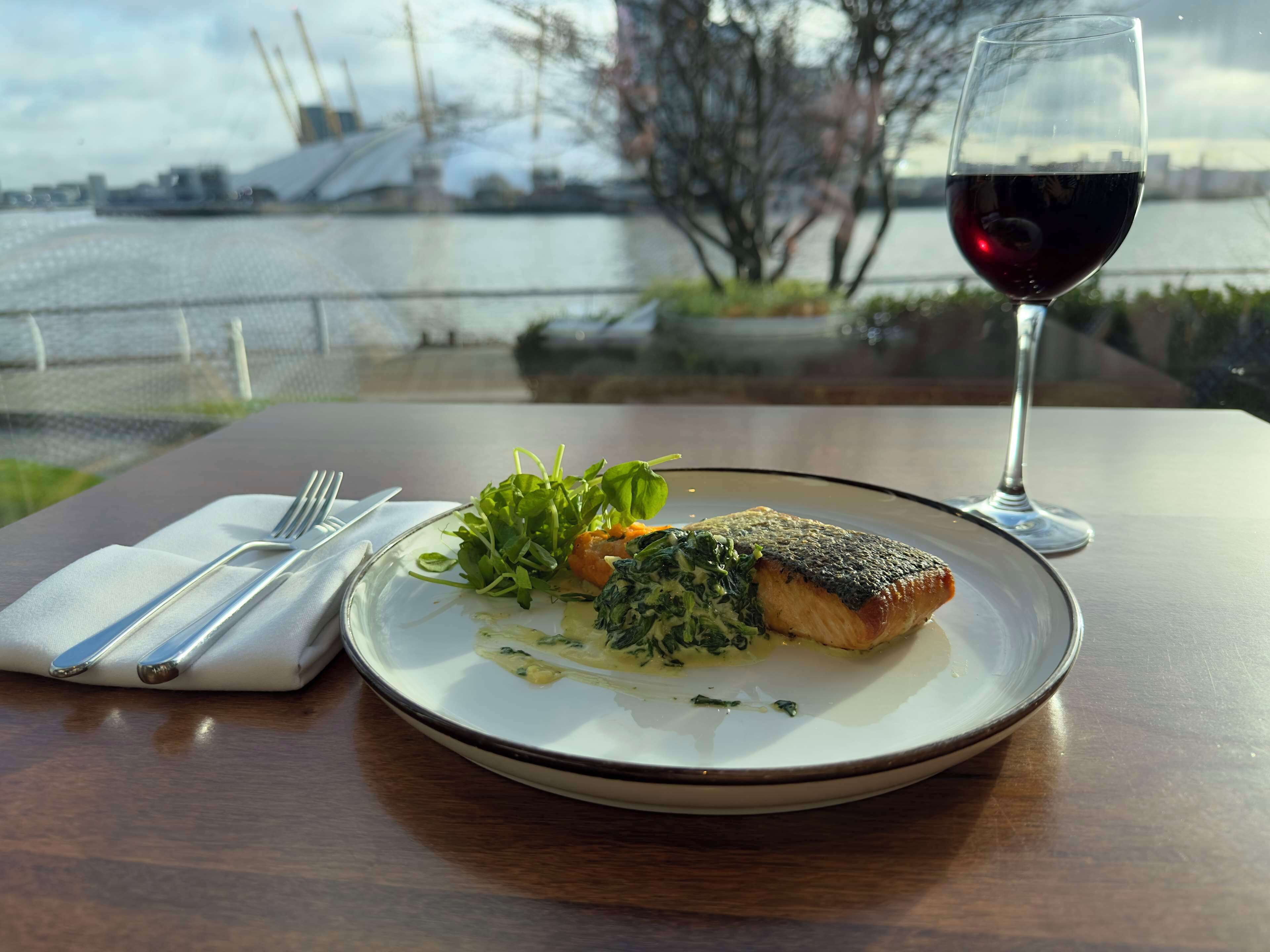 Une assiette de nourriture accompagnée d’un verre de vin rouge, posée sur une table d’où l’on aperçoit un plan d’eau et des bâtiments en arrière-plan.