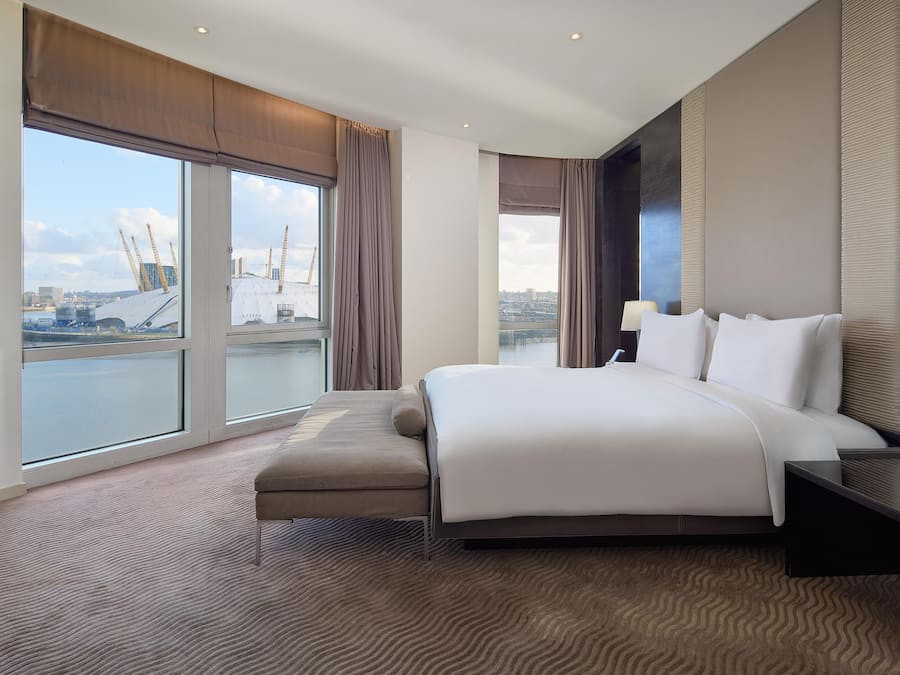 A modern hotel room with a large bed, a bench, and a view of a cityscape through large windows.