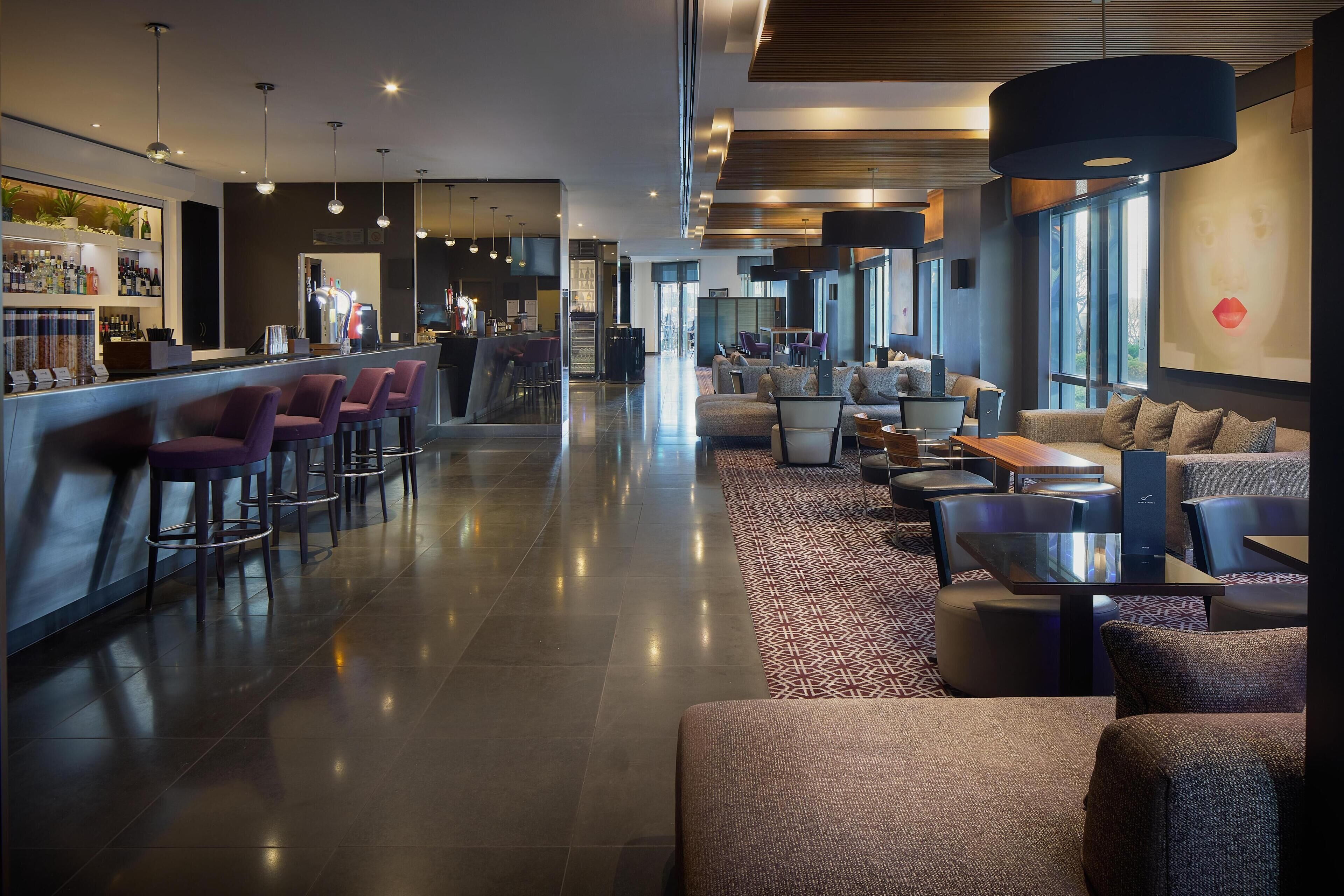 A modern hotel lobby with a bar area, seating, and a large window providing natural light.