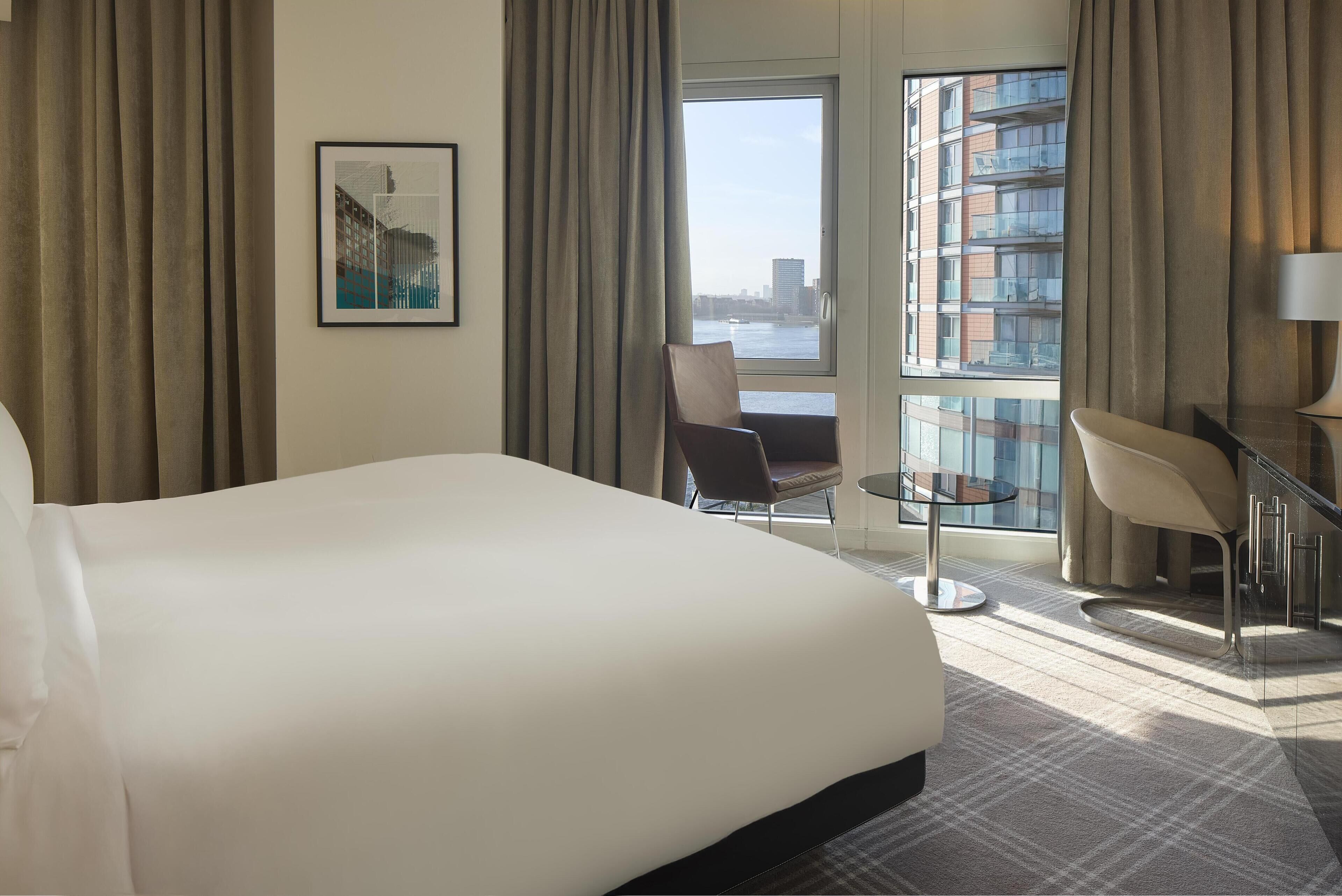 Ein Hotelzimmer mit einem großen Bett, einem Sessel, einem kleinen Tisch und Blick auf eine Stadtlandschaft durch ein Fenster.