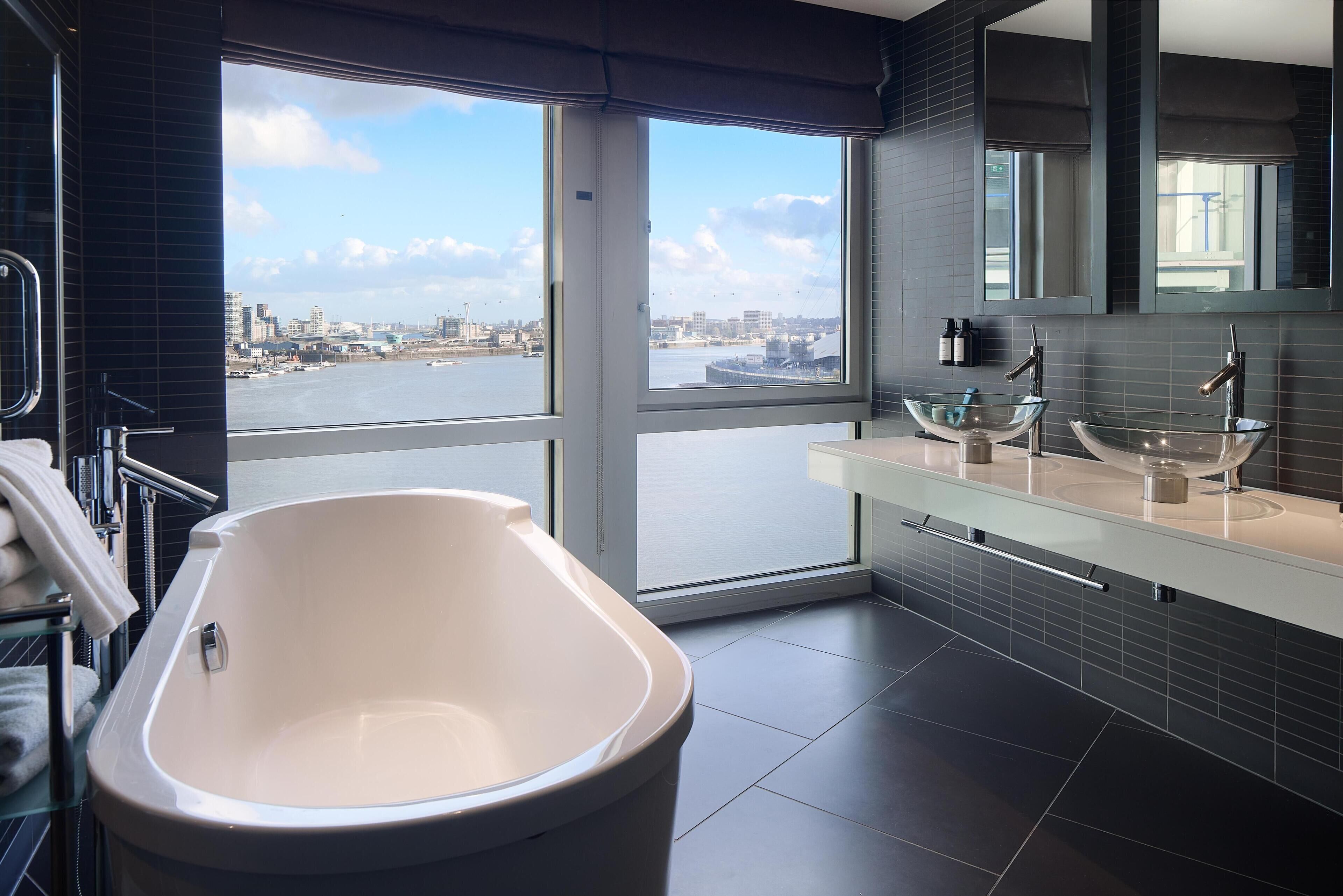 Une salle de bain moderne avec une grande baignoire, un lavabo en verre et une vue sur la ville grâce à de grandes fenêtres.