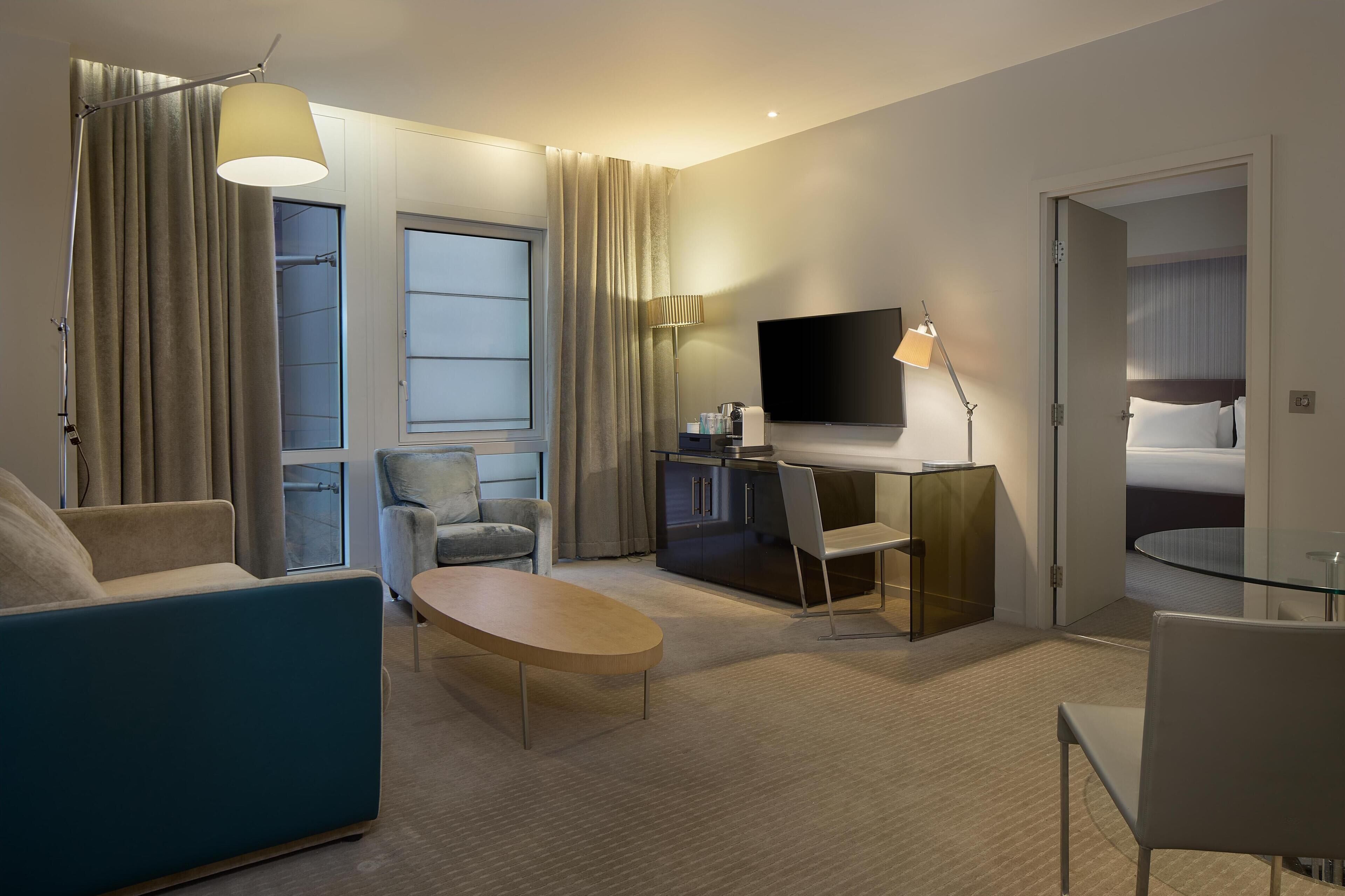Una habitación de hotel moderna con sofá, sillón, mesa de comedor, televisor y kitchenette.